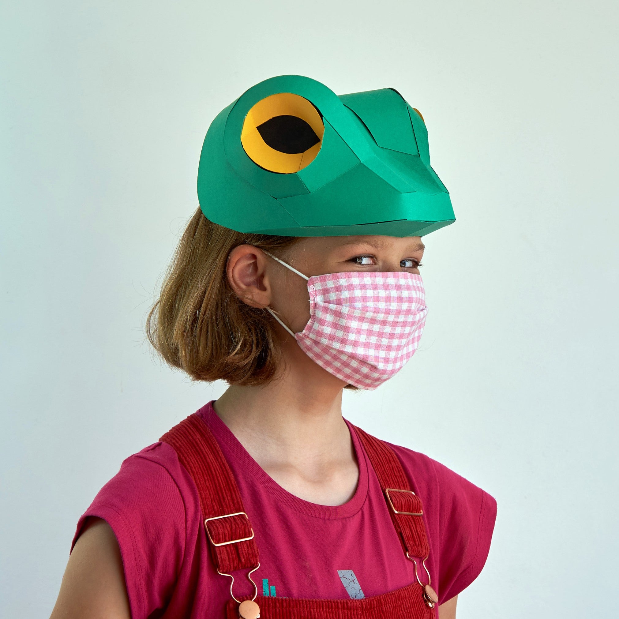 Child wearing a green frog hat and pink checkered mask on a light gray background