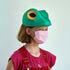 Child wearing a green frog hat and pink checkered mask on a light gray background