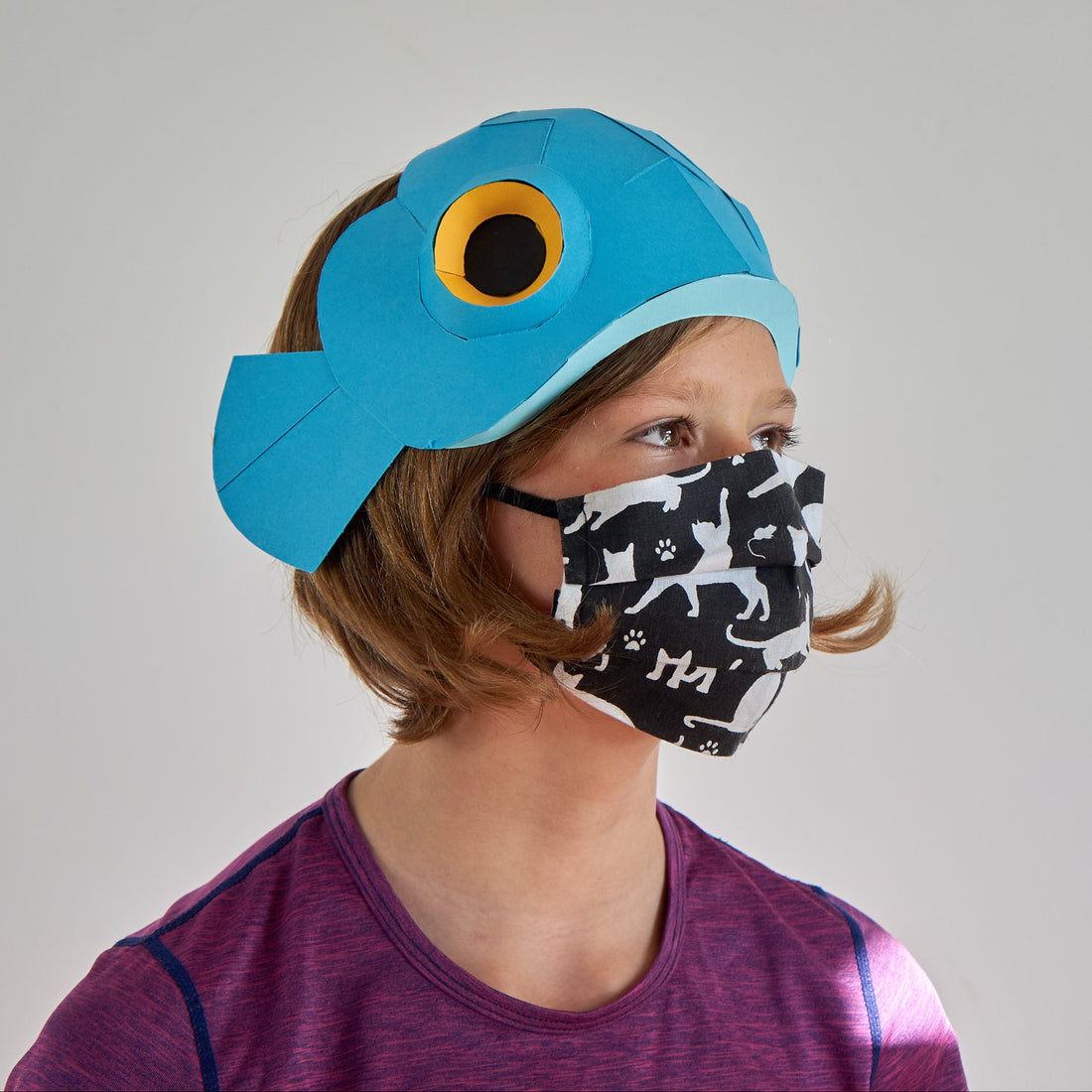 Person wearing a blue fish-shaped paper hat and black and white patterned face mask on a grey background
