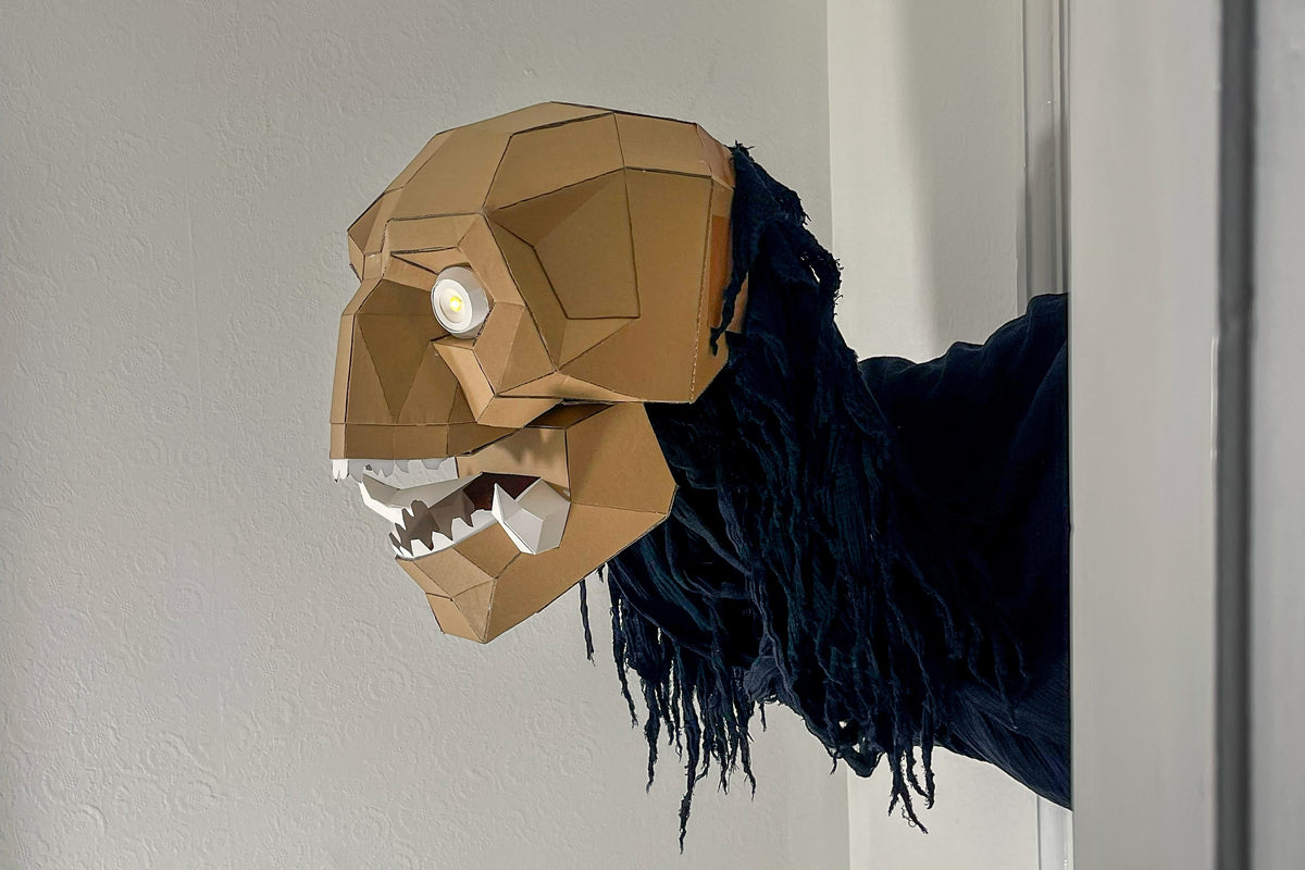 A stylized cardboard monster head with a single glowing white eye and open mouth showing white teeth, attached to a dark blue fringed neck, photographed against a plain light gray wall.