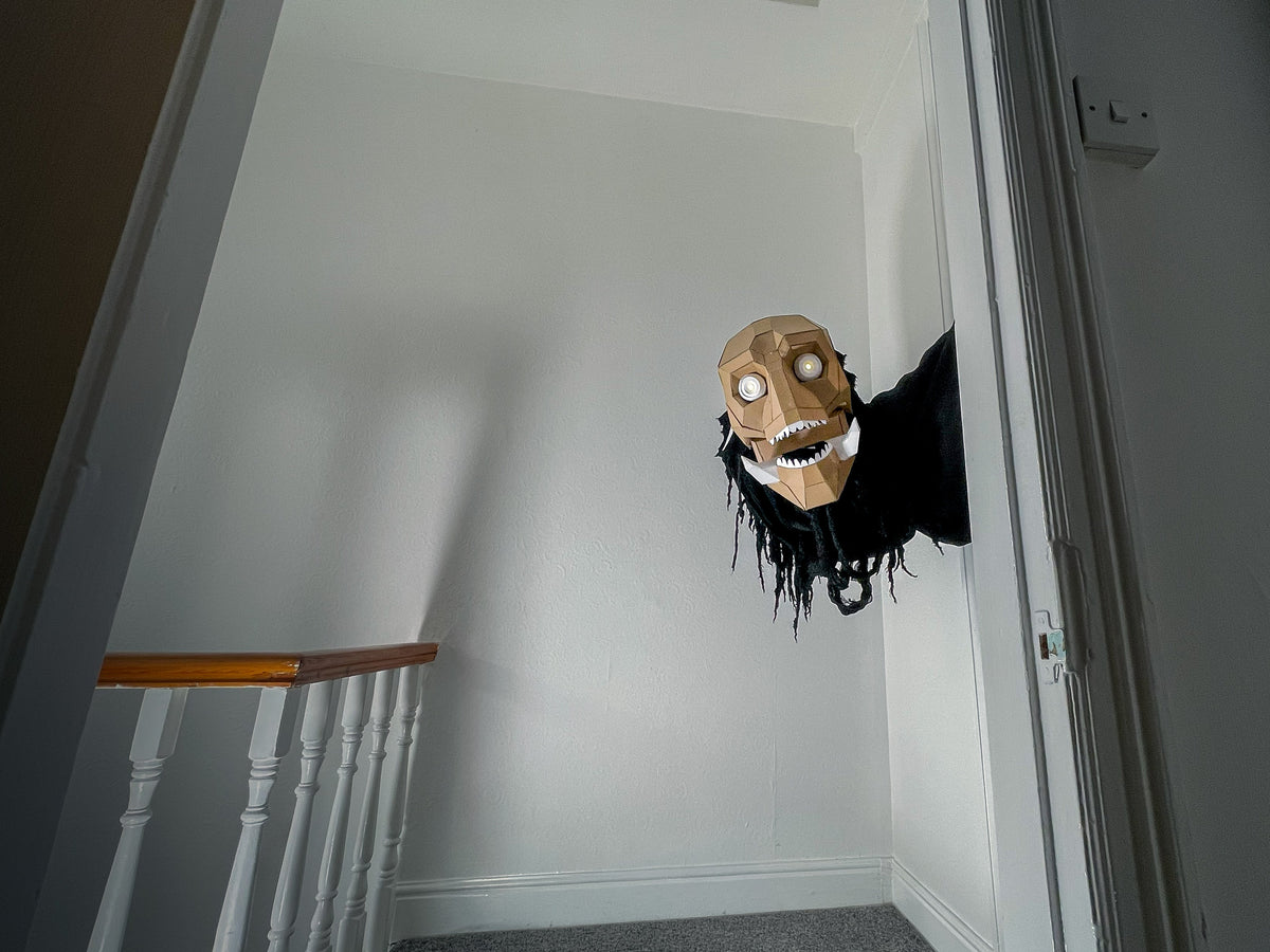 A creepy papercraft Halloween prop of a skull-faced figure with black tattered clothing peering out from a closed door in a residential hallway.
