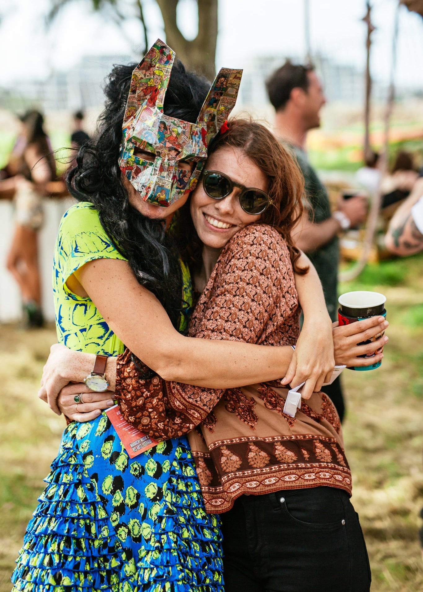 Two people hugging outdoors with one wearing a colorful mask and the other holding a cup.